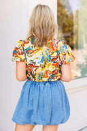 Woman wearing a colorful floral blouse and blue denim skirt standing in front of a white door with a brick wall background.