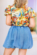 Woman wearing a colorful floral blouse and blue denim skirt standing in front of a white door with a brick wall background.