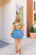 Woman wearing a colorful floral blouse and blue denim skirt standing in front of a white door with a brick wall background.