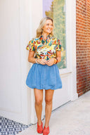 Woman wearing a colorful floral blouse and blue denim skirt standing in front of a white door with a brick wall background.