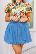 Woman wearing a colorful floral blouse and blue denim skirt standing in front of a white door with a brick wall background.