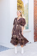 Woman wearing a brown floral dress standing in front of a white door with a brick. wall background.