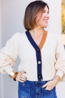 Woman wearing a cream cardigan with navy and brown trim accent and blue jeans.
