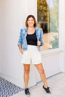 Woman wearing a denim jacket, black top, white corduroy shorts, and black sneakers standing in front of a white wall.