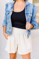 Person wearing a denim jacket, black top, and white corduroy shorts against a neutral background