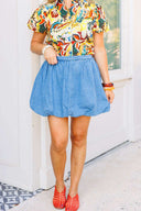 Woman wearing a colorful floral blouse and blue denim skirt standing in front of a white door with a brick wall background.
