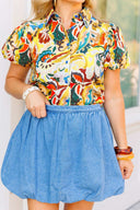 Woman wearing a colorful floral blouse and blue denim skirt standing in front of a white door with a brick wall background.