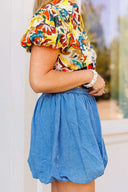 Woman wearing a colorful floral blouse and blue denim skirt standing in front of a white door with a brick wall background.