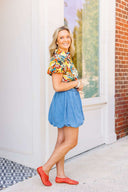 Woman wearing a colorful floral blouse and blue denim skirt standing in front of a white door with a brick wall background.
