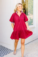 Woman wearing a crimson dress with a white wall and window in the background