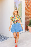 Woman wearing a colorful floral blouse and blue denim skirt standing in front of a white door with a brick wall background.