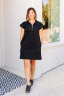 Woman wearing a black dress with gold accents standing in front of a white wall.