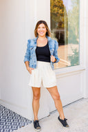 Woman wearing a denim jacket, black top, white corduroy shorts, and black sneakers standing in front of a white wall.