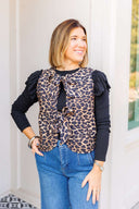 Woman wearing a leopard print vest with ruffled sleeve long sleeve underneath and blue jeans outdoors.