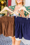 Woman holding two pairs of corduroy shorts - navy and brown - on hangers.