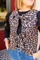 Woman wearing a leopard print vest with ruffled sleeve long sleeve underneath and blue jeans outdoors.