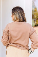 Woman wearing a beige sweatshirt with braided detail sleeves against a white wall.