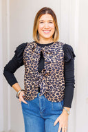 Woman wearing a leopard print vest with ruffled sleeve long sleeve underneath and blue jeans outdoors.