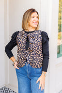 Woman wearing a leopard print vest with ruffled sleeve long sleeve underneath and blue jeans outdoors.