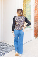 Woman wearing a leopard print vest with ruffled sleeve long sleeve underneath and blue jeans outdoors.
