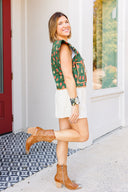 Woman standing outside boutique wearing a green floral vest, white shorts, and brown boots.
