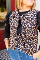 Woman wearing a leopard print vest with ruffled sleeve long sleeve underneath and blue jeans outdoors.