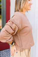 Woman wearing a brown sweatshirt with braided details, standing against a white wall.