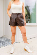 Person wearing brown leather shorts with studs and white ankle boots indoors.