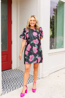 Woman wearing a floral dress with pink accents standing in front of a house entrance.