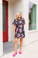 Woman in a floral dress standing outside a house with a red door.