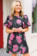 Woman wearing a black dress with pink floral pattern standing outdoors.