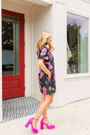 Woman in a floral dress and pink shoes standing outside a store with a red door.