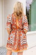 Woman wearing a patterned dress with orange accents, standing in a room with a window in the background.