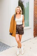Woman wearing a white tank top, brown leather shorts, and a tan jacket standing in front of a building.