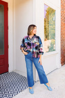 Woman wearing a colorful plaid shirt, blue jeans, and blue shoes standing in front of a white door.