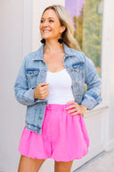 Woman wearing a denim jacket, white top, and pink shorts outdoors.